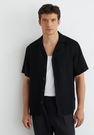 Hombre con camisa negra de manga corta desabrochada sobre camiseta blanca sin mangas, de pie frente a una pared blanca texturizada.