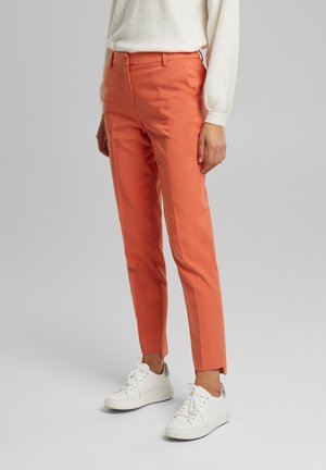 Personne portant un pantalon slim corail coupé à la cheville avec des fentes à l'avant, associé à des baskets blanches et un pull crème à manches longues.