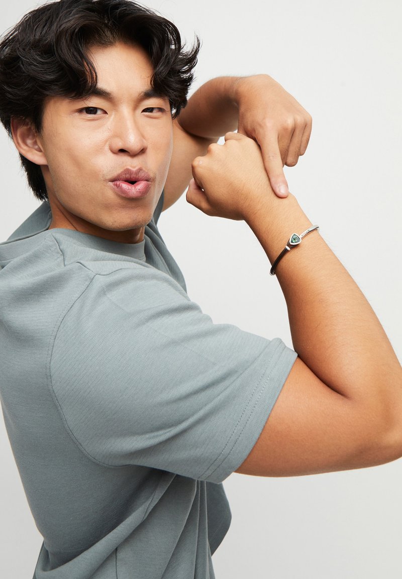 Pulsera de plata con una piedra triangular verde, llevada en una muñeca musculosa. El portador lleva una camisa de manga corta gris y señala la pulsera.