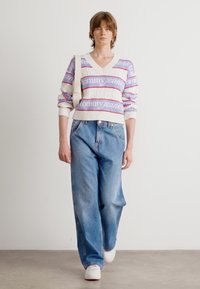 Witte v-hals trui met rood en blauwe horizontale strepen, voorzien van de tekst "tommy jeans", gecombineerd met lichtblauwe losse spijkerbroeken.