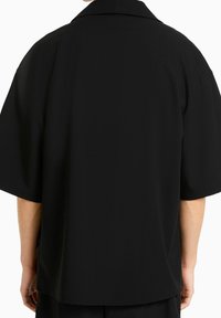 Musta lyhythihainen paita löysällä leikkauksella, jossa on klassinen kaulus ja pehmeä kangas. Ei näkyviä kuvioita tai kiinnikkeitä.