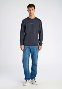 Mörkblå sweatshirt med rund halsringning och ribbade ärmslut, med en ljusblå "LINDBERGH"-logotyp. Matchas med ljusblå jeans och vita sneakers.