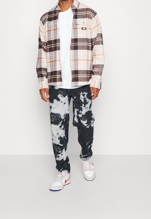 Uomo con camicia a quadri marrone e beige sopra una t-shirt bianca, jeans tie-dye nero e bianco e sneakers bianche con logo rosso.