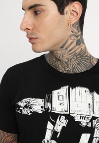 Schwarzes Baumwoll-T-Shirt mit einem weißen grafischen Design eines AT-AT-Walkers, der von einem Piloten besetzt ist, mit einem klassischen Rundhalsausschnitt.