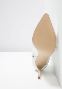 Semelle beige claire d'une chaussure blanche à talon haut et bout pointu, reposant verticalement contre un coin blanc.