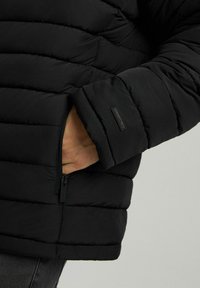 Czarna kurtka puchowa z poziomymi segmentami pikowania, z zamykaną na zamek kieszenią na rękawie oraz metką z logo w pobliżu mankietu.