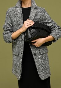 Blazer gris texturizado con un bolsillo tipo parche, llevado sobre un vestido negro. Sosteniendo un bolso tipo clutch negro, acolchado, con la parte superior fruncida.