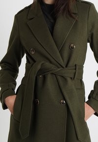 Doppelreihiger olivgrüner Mantel mit tailliertem Gürtel, aus strukturiertem Stoff. Verfügt über große braune Knöpfe und strukturierte Revers.