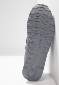 Semelle d'une chaussure New Balance grise montrant un motif détaillé de la semelle et un logo, positionnée verticalement contre un mur blanc.