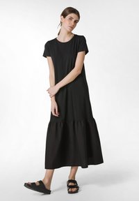 Černé midi šaty s krátkými rukávy, s volánkovým lemem, vyrobené z měkkého materiálu. Modelka má na sobě černé sandály.