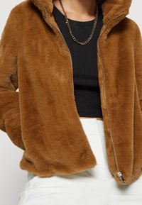 Chaqueta de piel sintética en marrón con cierre frontal de cremallera y capucha. Presenta una textura suave y un corte holgado, combinada con una camiseta negra de canalé debajo.