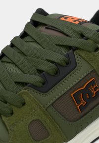 Green sneaker featuring a mix of textured mesh and suede materials, flat laces, a black accent, and an orange logo on the side.