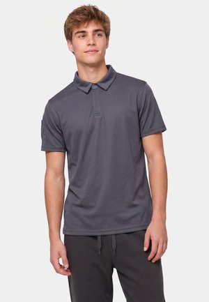 Giovane uomo con polo a maniche corte grigio scuro e pantaloni scuri, in piedi e che guarda leggermente di lato con un'espressione neutra.
