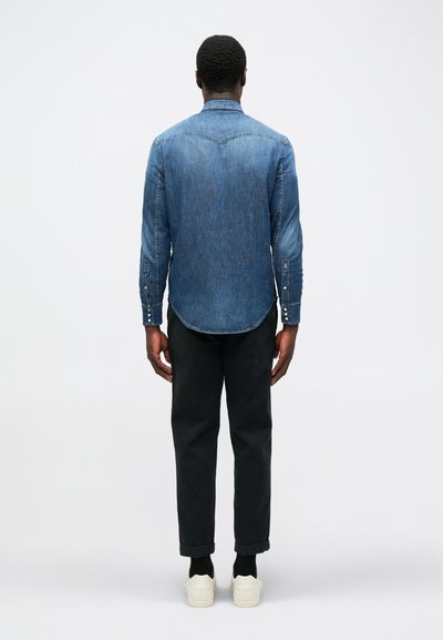 Camicia di jeans di colore blu medio, con dettagli a bottoni sulle maniche, orlo curvo, indossata con pantaloni scuri e scarpe bianche, vista da dietro.