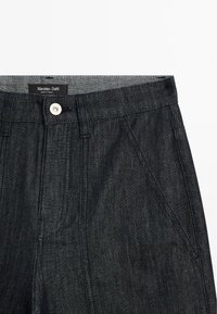 Tmavé džíny s knoflíkovým zapínáním, s nášivkou na pase. Látka má hladký, mírně strukturovaný povrch.