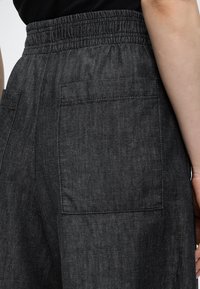 Gros plan sur un pantalon gris foncé avec une taille élastique et une grande poche arrière côté droit, porté avec un haut noir.