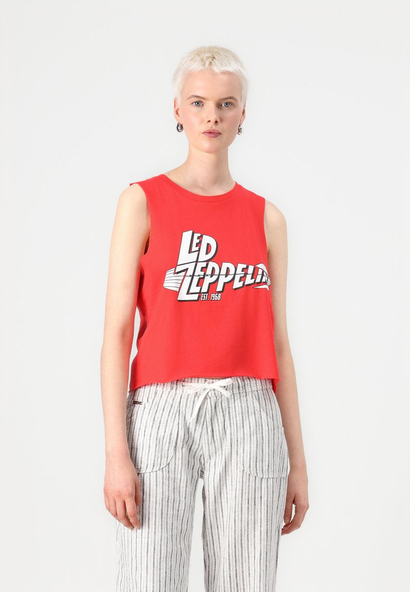Red sleeveless crop top featuring a Led Zeppelin graphic in white and black. The model pairs it with light striped fabric pants.