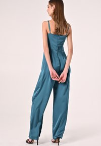 Tuta di raso teal con un corpetto aderente, spalline sottili e pantaloni a gamba larga. Dettagli notevoli includono una chiusura con zip e una lucentezza delicata.