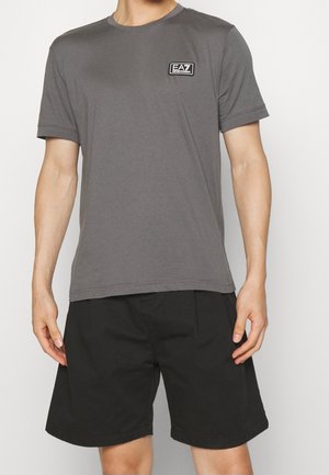 Homme portant un T-shirt gris à manches courtes avec le logo EA7 sur la poitrine et un short noir longueur genou, debout devant un fond uni.