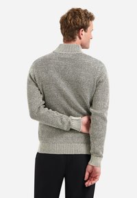 Gebreide trui in lichtgrijs met een textuurpatroon en contrasterende ribbels op de kraag, manchetten en zoom. Model heeft de handen achter de rug gevouwen.