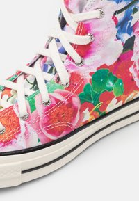 Wysokie trampki z białymi sznurowadłami, metalowymi oczkami i materiałem z płótna, zdobione kolorowym wzorem kwiatowym z zielonymi liśćmi.