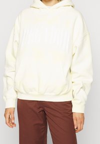 Sudadera color crema con capucha, que presenta un gran texto blanco "PEGADOR", un bolsillo frontal tipo canguro y puños y dobladillo de canalé.