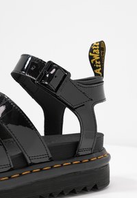 Sandale en cuir verni noir avec bride à boucle réglable, semelle épaisse avec surpiqûres jaunes et languette en tissu brandée au talon.