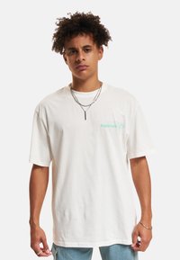 Weißes Baumwoll-T-Shirt mit lockerer Passform, kurzen Ärmeln und einem hellgrünen Logo "Double Espresso" auf der Brust. Tragen mit geschichteten silbernen Halsketten.