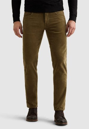 Homme portant un pantalon en velours côtelé marron, une chemise noire à manches longues et des bottes marron foncé à lacets, debout devant un fond uni.