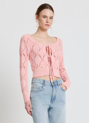 Donna che indossa un cardigan rosa lavorato a maglia con motivi floreali e lacci frontali, abbinato a jeans chiari a vita alta e orecchini floreali.