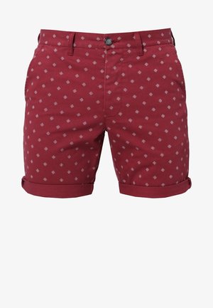 Hombre con camisa azul abotonada sobre una camiseta blanca, pantalones cortos rojos con estampado y zapatos claros tipo slip-on, de pie frente a un fondo liso.