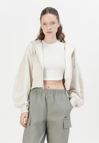 Korte beige hoodie met een ritssluiting aan de voorkant, gecombineerd met een lichtgrijze getailleerde tanktop en groene cargo broek met zijzakken.