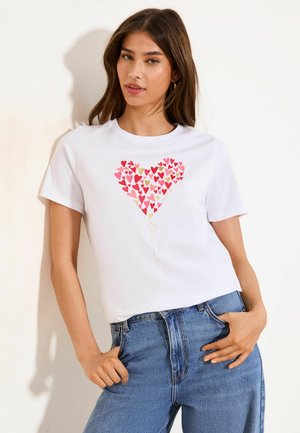 Fiatal nő fehér pólóban, melyen többszínű szív alakú léggömb mintája van, és kék farmerben, egyszínű háttér előtt áll.