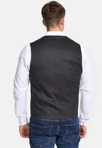 Dunkelgraue Weste mit strukturiertem Muster, Knopfverschluss und tailliertem Schnitt, kombiniert mit einem weißen Langarmshirt und blauen Jeans.
