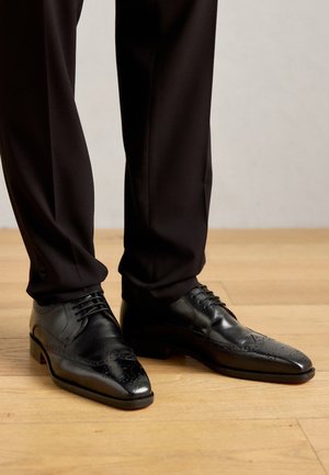 Hombre con zapatos de vestir de cuero negro pulido y pantalones negros de pie sobre un suelo de madera clara.