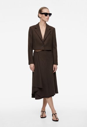 Vrouw met een donkerbruine cropped blazer, asymmetrische midi-rok, zwarte zonnebril en zwarte platte sandalen met bandjes, staand op een witte achtergrond.