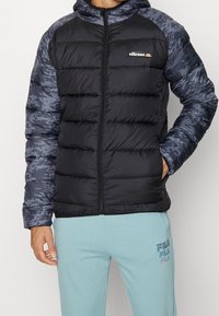 Puffjacka med svart front, marinblå pixelerat mönster på ärmar och huva. Fram dragkedja, lättviktig och med kviltad textur. Märkeslogotyp på bröstet.