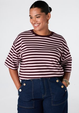 Lachende vrouw die een paars-roze gestreept T-shirt met halve mouwen en hoog getailleerde donkerblauwe spijkerbroek met gouden knopen draagt.