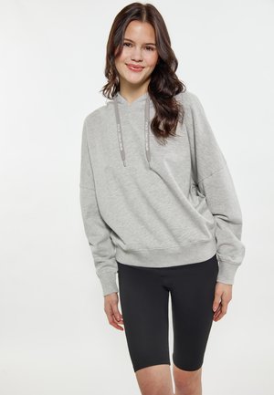 Jonge vrouw met lang donker haar, gekleed in een lichtgrijze hoodie en zwarte knielange shorts, staand tegen een effen witte achtergrond.