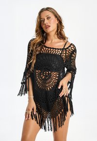 Czarna narzutka szydełkowa o luźnym, ażurowym wzorze. Posiada falowany brzeg oraz długie frędzle na rękawach, prezentujące misternie wykonane wzory.