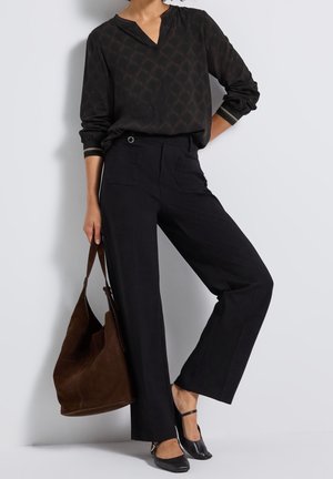 Persoon draagt een zwarte top met lange mouwen en patroon, zwarte wijde broek, zwarte Mary Jane schoenen, houdt een grote bruine suède schoudertas vast.