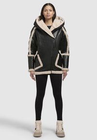 Sort læderjakke med beige skindfor, oversized hætte, fremtrædende syninger og vinklede lommeakcenter. Båret med sorte leggings og beige støvler.