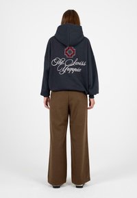 Marineblauwe hoodie met een grote bordeauxrode kruisgrafiek en de tekst "The Swiss Yuppie" op de achterkant; gecombineerd met bruine wijde broek.