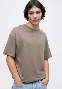Young person with medium-length dark hair wearing a loose taupe T-shirt and light blue jeans, hands in pockets, looking slightly right.