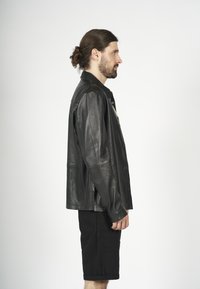 Giacca di pelle nera con un design a colletto, chiusura a bottoni e spacchi laterali. Il modello indossa pantaloni corti neri, in piedi su uno sfondo bianco.