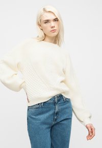 Ξανθιά γυναίκα που φοράει κρεμ πουλόβερ πλεκτό με κοτσίδες και μπλε τζιν, καθώς στέκεται με το ένα χέρι στη μέση ενάντια σε απλό λευκό φόντο.