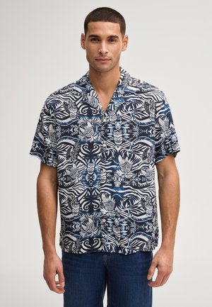 Mann mit kurzen Haaren, der ein blau-weiß gemustertes kurzärmeliges Hemd mit Knöpfen und dunkle Jeans trägt, vor einem schlichten Hintergrund stehend.