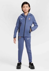 Dziecko stoi w niebieskim dresie sportowym Paris Saint-Germain, w czarnych sneakersach, z rękami w kieszeniach na jednolitym tle.