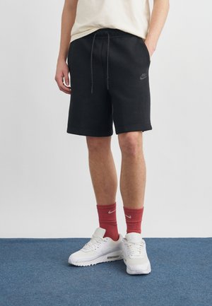 Muž oblečený v čiernych športových kraťasoch Nike, červených ponožkách Nike a bielych teniskách Nike, stojaci na modrom koberci pred jednoduchým svetlým pozadím.