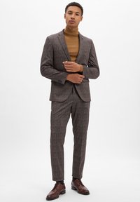 Brauner karierter Anzug für Herren mit einem taillierten Blazer und passender Hose, kombiniert mit einem braunen Rollkragenpullover und dunkelbraunen Schuhen.
